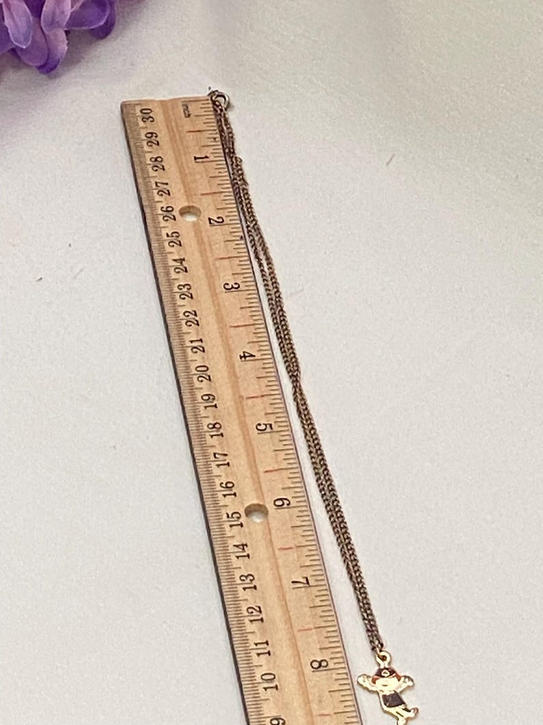 May include: A gold-toned necklace with a pendant featuring a cartoon figure. The necklace is measured with a wooden ruler, showing the chain's length. The pendant is a small, detailed design with red and black accents.
