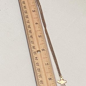 May include: A gold-toned necklace with a small pendant in the shape of a figure with outstretched arms. The pendant has red, pink, and white accents. The necklace is next to a wooden ruler with inch markings.