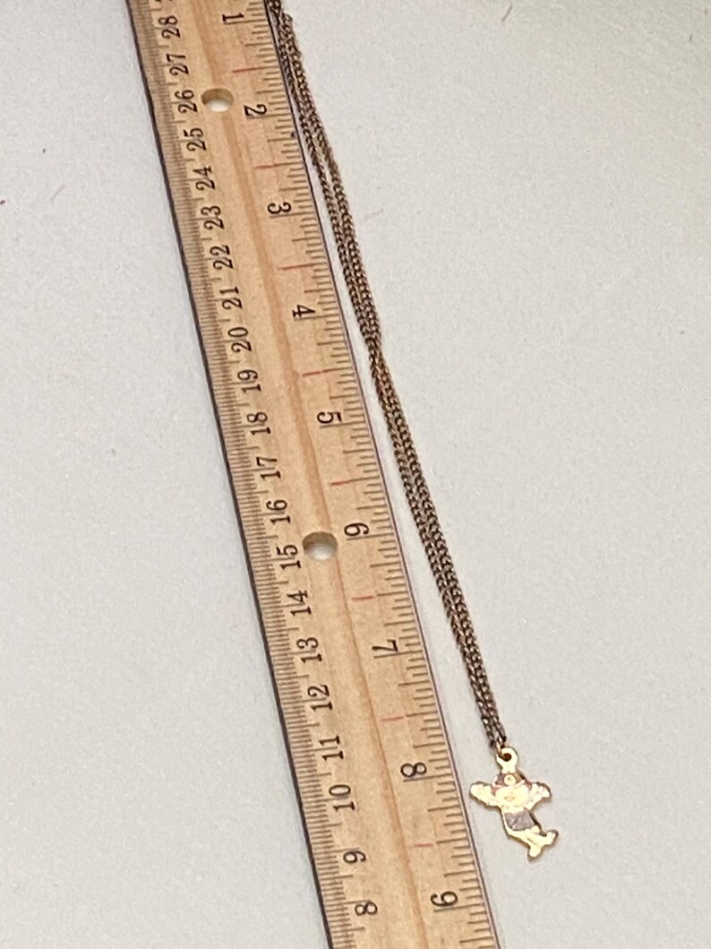 May include: A gold-toned necklace with a small pendant in the shape of a figure with outstretched arms. The pendant has red, pink, and white accents. The necklace is next to a wooden ruler with inch markings.