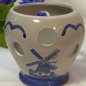 May include: A cream-colored ceramic candle holder with a blue windmill design and wave accents. The holder has circular cutouts and a blue base. The windmill illustration is detailed, and the overall design is reminiscent of Dutch pottery.