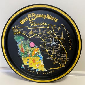 May include: A black metal tray with a yellow rim, featuring a map of Florida with Disney characters. The tray has the text "Walt Disney World Florida" and "GULF OF MEXICO" printed on it.
