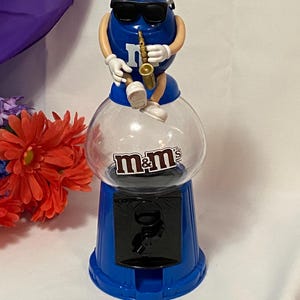 May include: A blue M&M candy dispenser with a clear globe. The M&M character on top wears sunglasses and plays a saxophone. The dispenser has the M&M's logo and a coin slot.