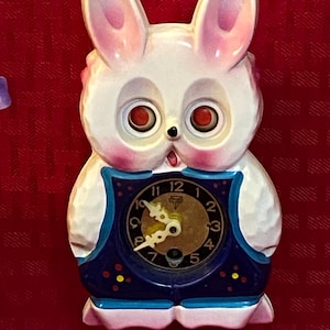May include: A vintage bunny-shaped clock with a white body, pink ears, and red eyes. The clock face is gold with black numbers and hands, set within a blue frame. A small pendulum hangs below.