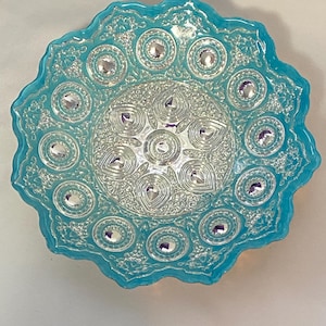 May include: A decorative, turquoise-colored glass plate with a scalloped edge. The plate features a detailed, silver-colored, ornate design with circular and teardrop-shaped accents. The center of the plate has a similar design, with a series of concentric circles.