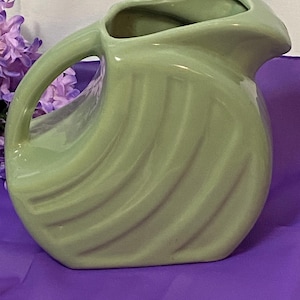 May include: A light green ceramic pitcher with a curved handle and a spout. The pitcher has a rounded body with vertical, wave-like ridges. The pitcher is on a purple surface.