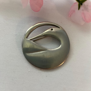 May include: A silver-toned brooch with a bird design. The brooch is circular, with a stylized bird silhouette. The bird's head and beak are prominent, and the body is a curved shape. The brooch is set against a white background with pink flowers.