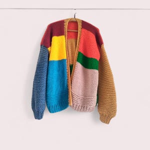 Può includere: Un cardigan colorato e a maglia grossa con un design patchwork. Il maglione presenta pannelli blu, gialli, rossi, rosa e marroni. Le maniche sono lunghe e il cardigan è aperto sul davanti, appeso a una gruccia di legno.