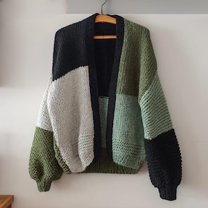 Puede incluir: Un cárdigan de punto grueso con un diseño de retazos. El patrón de bloques de color presenta negro, gris y tonos de verde. El suéter abierto está colgado en una percha de madera contra una pared blanca.