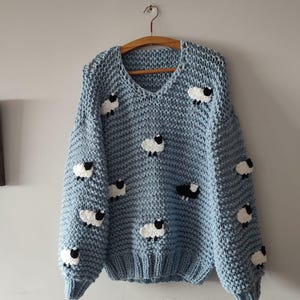 May include: A light blue knit sweater with a V-neck and long sleeves. The sweater is adorned with multiple white and black sheep appliqués. The garment is hanging on a wooden hanger against a light gray wall.