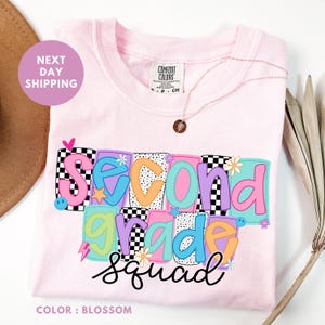 A pink t-shirt with the text "Second Grade Squad" printed on it, along with colorful geometric shapes and flowers. The t-shirt is displayed on a white background, and there is a brown hat and some leaves in the background.