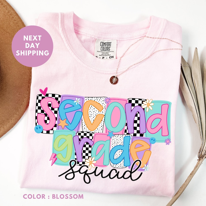 A pink t-shirt with the text "Second Grade Squad" printed on it, along with colorful geometric shapes and flowers. The t-shirt is displayed on a white background, and there is a brown hat and some leaves in the background.