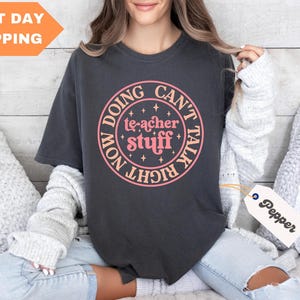 May include: A dark grey t-shirt with a pink and white graphic that says "Doing Teacher Stuff Now Can't Talk Right Now".
