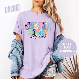 A woman wearing a purple t-shirt with a colorful graphic design and text, standing in front of a white background.