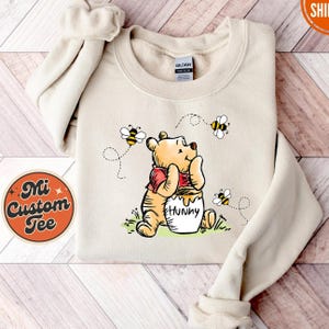 May include: A beige sweatshirt with a cartoon image of Winnie the Pooh holding a pot of honey. The image is surrounded by bees and the text "Hunny" is printed on the pot.