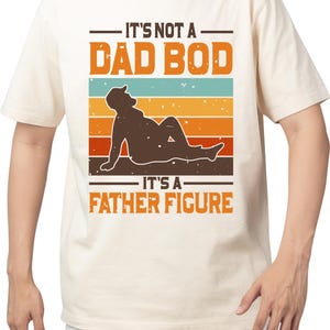 May include: Cream-colored t-shirt with a graphic design. The design features the text "IT'S NOT A DAD BOD IT'S A FATHER FIGURE" above and below a silhouette of a relaxed person. The background has horizontal stripes in orange, blue, and brown.