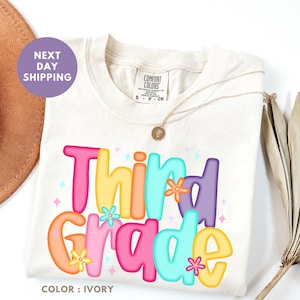 a white t-shirt with a colorful graphic design that says "Third Grade" in a playful font.