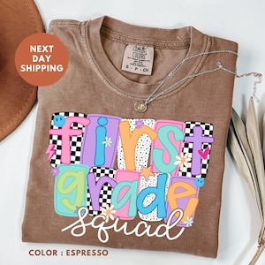 a brown t-shirt with a colorful design that says "FIRST GRADE SQUAD" in a playful font. The t-shirt is placed on a white surface, and there is a brown hat and some leaves in the background.