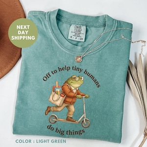 Może przedstawiać: Jasnozielona koszulka z napisem "Off to help tiny humans do big things" i ilustracją żaby na skuterze. Żaba nosi okulary i kurtkę, niosąc torbę z przyborami szkolnymi. Srebrny naszyjnik jest zarzucony na koszulkę.