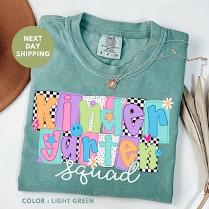 May include: Light green t-shirt with the words "Kindergarten Squad" in colorful, block letters. The shirt has a crew neck and short sleeves. A gold necklace with a pendant is draped across the shirt.