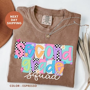 a brown t-shirt with a colorful design that says "Second Grade Squad" printed on it. The t-shirt is placed on a white surface, and there is a brown hat and some leaves nearby.