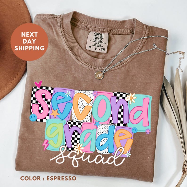 a brown t-shirt with a colorful design that says "Second Grade Squad" printed on it. The t-shirt is placed on a white surface, and there is a brown hat and some leaves nearby.