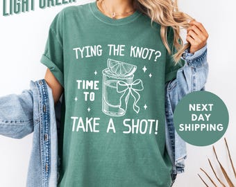 Puede incluir: Camiseta verde claro con texto blanco que dice "TYING THE KNOT? TIME TO TAKE A SHOT!" y un gráfico de un vaso de chupito con una rodaja de lima y un lazo.