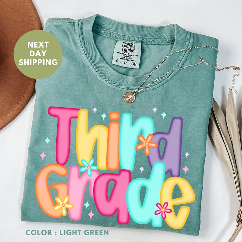 A green t-shirt with the text "Third Grade" printed on it, along with colorful flowers and stars. The t-shirt is displayed on a white background, and there is a brown hat and some leaves in the background.