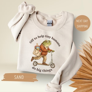 May include: Sand-colored sweatshirt with a graphic of a frog riding a scooter, carrying a tote bag filled with school supplies. Text reads "Off to help tiny humans do big things."