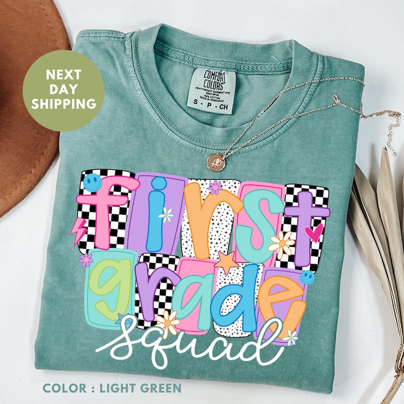 A green t-shirt with colorful text that says "FIRST GRADE SQUAD" printed on it, along with a necklace and some decorative leaves.