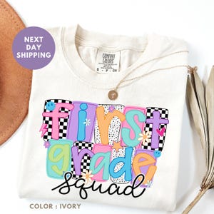 a white t-shirt with a colorful graphic design that says "FIRST GRADE SQUAD" in a playful font.