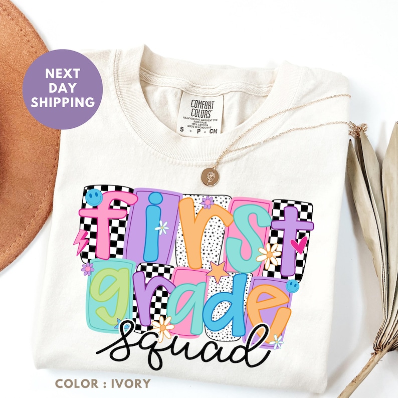 a white t-shirt with a colorful graphic design that says "FIRST GRADE SQUAD" in a playful font.