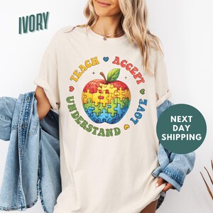 Könnte beinhalten: Ein T-Shirt in Elfenbein mit einem Grafikdruck einer Regenbogen-Puzzle-Apfel mit dem Text "Teach Accept Understand Love" drumherum.