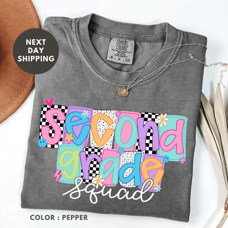 a gray t-shirt with colorful text that says "Second Grade Squad" printed on it. The t-shirt is folded and placed on a white surface, with a brown hat and some flowers nearby.