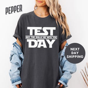 May include: A dark gray t-shirt with white text that reads "TEST MAY THE SKILLS BE WITH YOU DAY".