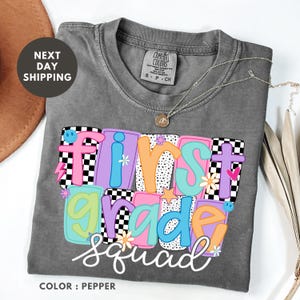 a gray t-shirt with a colorful design that says "First Grade Squad" in a playful font.