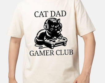 May include: Cream-coloured t-shirt with a black graphic of a cat wearing headphones and holding a game controller. The text "CAT DAD" is above the cat, and "GAMER CLUB" is below.