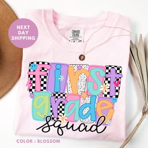 A pink t-shirt with a colorful graphic design that says "FIRST GRADE SQUAD" and features various colorful letters and shapes.