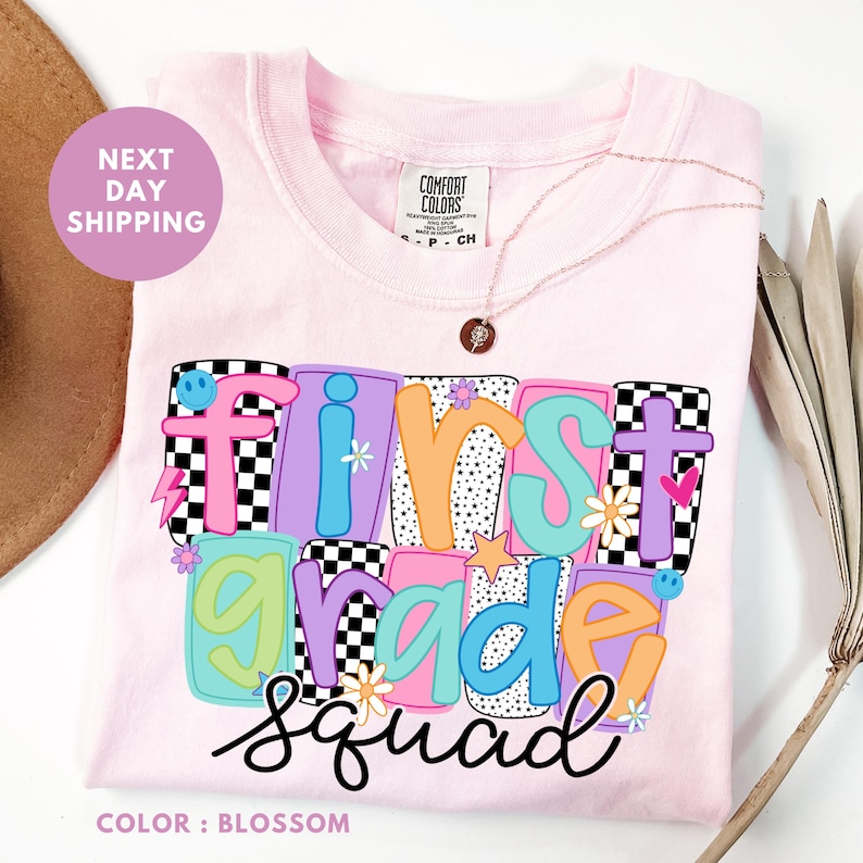 A pink t-shirt with a colorful graphic design that says "FIRST GRADE SQUAD" and features various colorful letters and shapes.