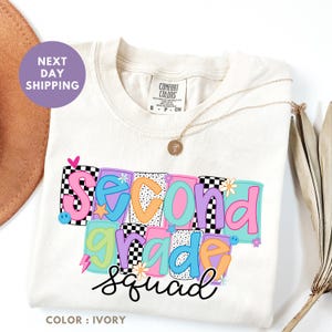 a white t-shirt with a colorful graphic design that says "Second Grade Squad" in bold letters. The t-shirt is placed on a white background, and there is a brown hat and a plant visible in the background.