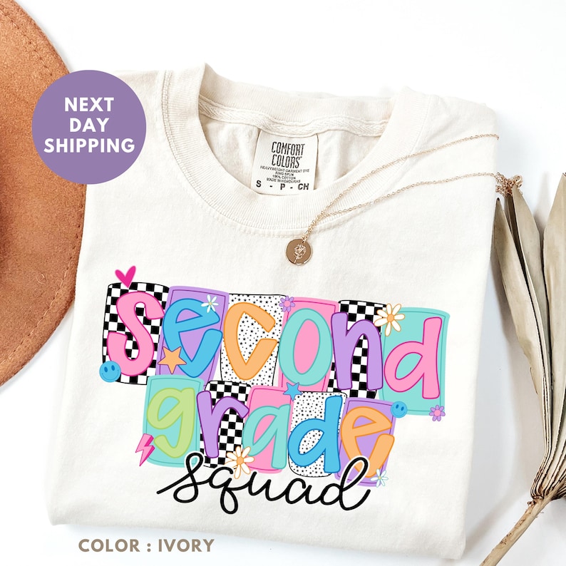 a white t-shirt with a colorful graphic design that says "Second Grade Squad" in bold letters. The t-shirt is placed on a white background, and there is a brown hat and a plant visible in the background.