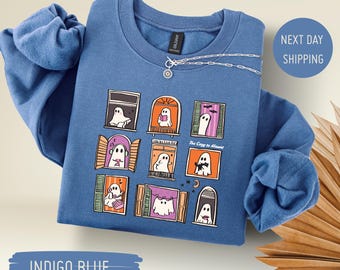 Puede incluir: Sudadera azul índigo con un diseño caprichoso de fantasmas en ventanas. Incluye el texto "Too Cozy to Haunt". Un collar plateado con un colgante está sobre la sudadera. Las mangas son visibles.
