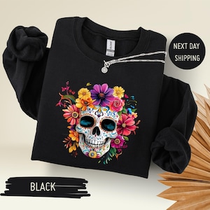 May include: Black sweatshirt featuring a colorful sugar skull design with vibrant flowers. The skull has intricate patterns and bright blue eye sockets. A silver necklace with a pendant rests on the sweatshirt. The text "BLACK" is at the bottom.