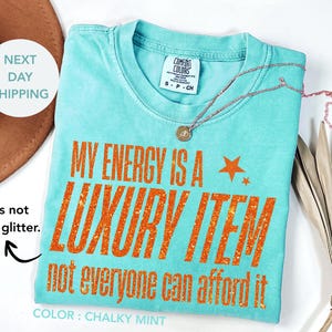 Puede incluir: Una camiseta turquesa con la frase "MY ENERGY IS A LUXURY ITEM not everyone can afford it" en letras de purpurina naranja. La camiseta tiene cuello redondo y una pequeña etiqueta. También se ve un collar dorado y una hoja de palma seca.