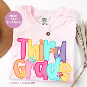 A pink t-shirt with the text "Third Grade" printed on it, surrounded by colorful flowers and stars.