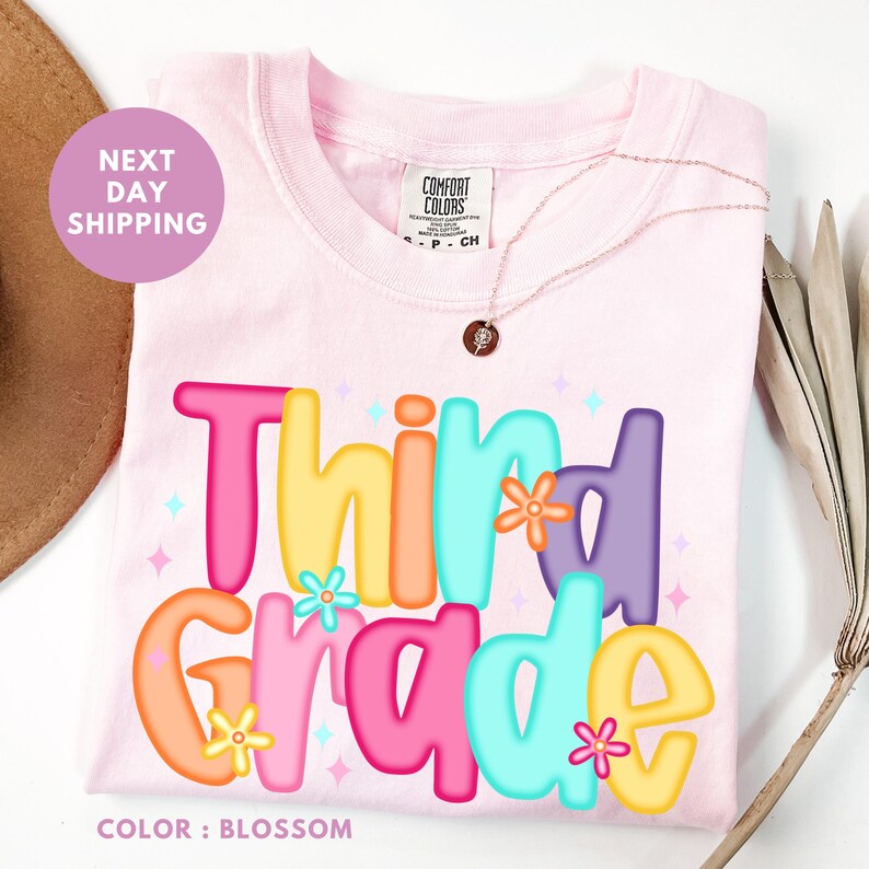 A pink t-shirt with the text "Third Grade" printed on it, surrounded by colorful flowers and stars.