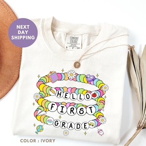 以下が含まれることがあります： 鮮やかな「Hello First Grade」のデザインが施されたアイボリー色のTシャツ。学校をテーマにしたアイコンとデイジーの花がブレスレットのような模様になっています。Tシャツはクルーネックで半袖です。金のネックレスがTシャツにかけられています。