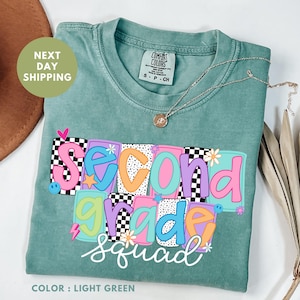 Puede incluir: Camiseta verde claro con la frase "Second Grade Squad" en un diseño colorido. El diseño incorpora elementos de tablero de ajedrez y florales. Un collar plateado está sobre la camiseta. Marca Comfort Colors.