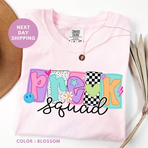 May include: A light pink t-shirt with the words "Pre K Squad" in a colorful, playful design. The design includes a smiley face, stars, flowers, and a checkered pattern. A gold necklace with a pendant is draped on the shirt.