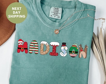 Pode incluir: Camiseta verde claro com o nome "MADISON" em letras com tema natalino. As letras são decoradas com Papai Noel, listras, glitter, neve, um boneco de neve, uma coroa e renas. Um colar de prata está drapeado sobre a camisa.