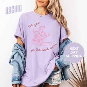 May include: A lavender t-shirt with a pink graphic of a dog surfing on a wave. The text "see you on the next wave" is printed on the shirt.